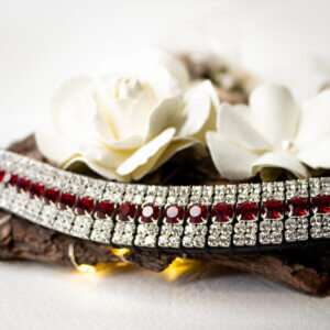 A handmade red browband with red and clear crystals on a white background with cream flowers and a piece of tree bark